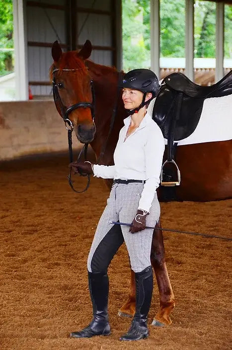 Woman in equestrian attire standing next to a horse in an indoor arena.